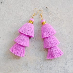 May include: A pair of pink tassel earrings with gold accents. The earrings are layered with three tiers of tassels.