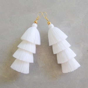May include: A pair of white tassel earrings with gold hooks. The tassels are layered and have a tiered look.
