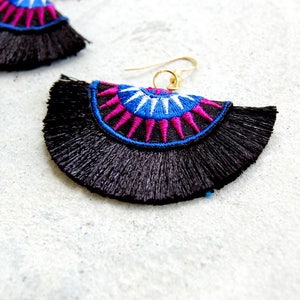 May include: A pair of black tassel earrings with a colorful geometric design. The earrings feature a blue, white, and pink pattern with a gold hook.