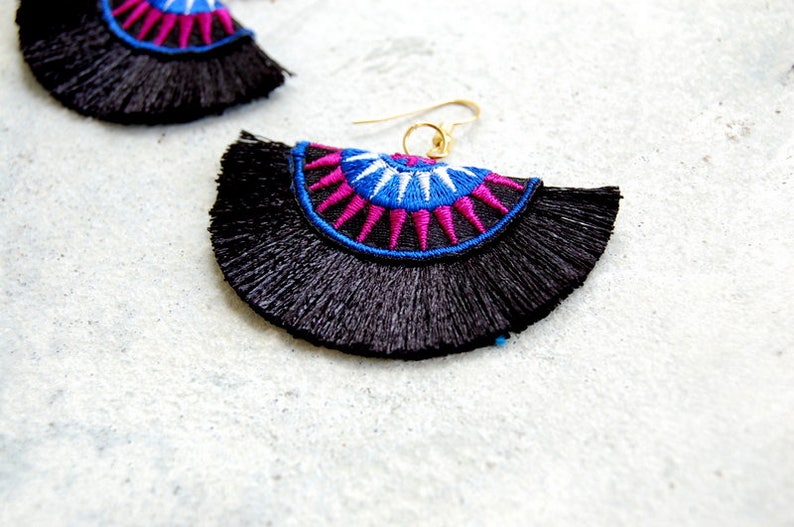 May include: A pair of black tassel earrings with a colorful geometric design. The earrings feature a blue, white, and pink pattern with a gold hook.
