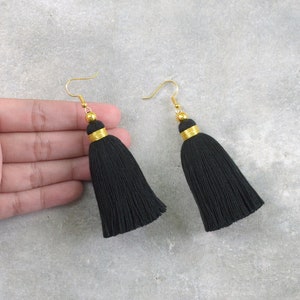 Puede incluir: Un par de pendientes de borla negra con detalles dorados. Los pendientes tienen un cierre de gancho simple.