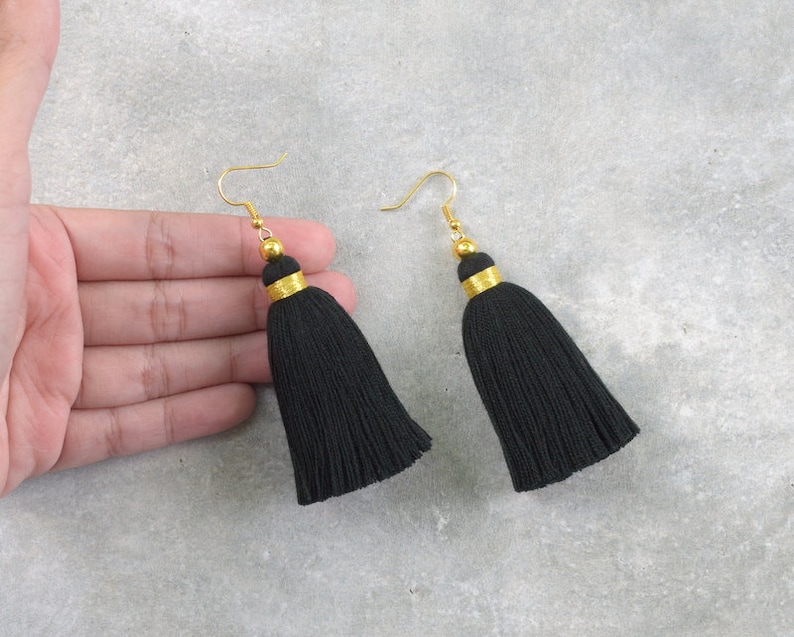 Puede incluir: Un par de pendientes de borla negra con detalles dorados. Los pendientes tienen un cierre de gancho simple.