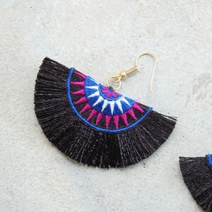 May include: Black tassel earrings with a colorful embroidered design. The design features a circle with pink, blue, and white accents.