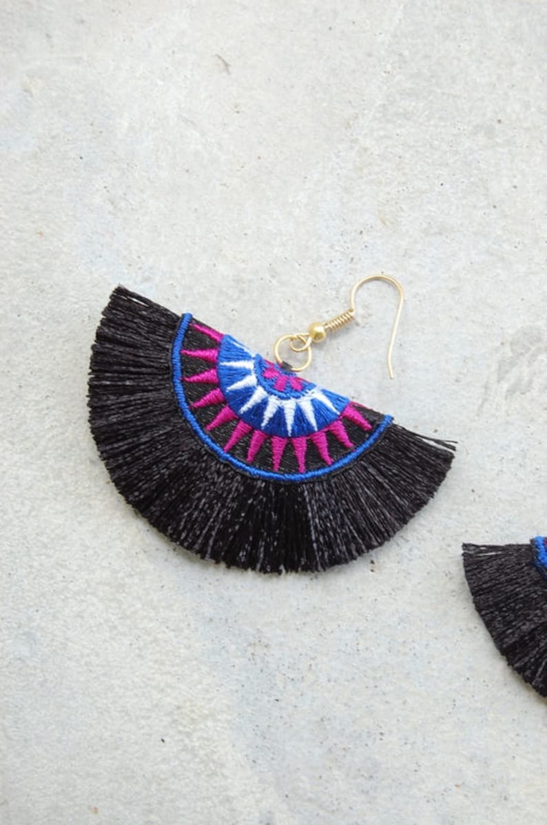 May include: Black tassel earrings with a colorful embroidered design. The design features a circle with pink, blue, and white accents.
