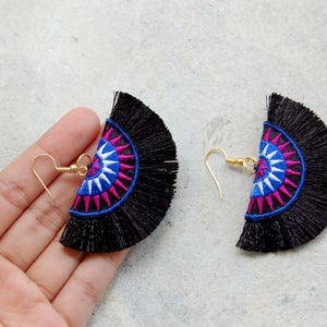 May include: A pair of black tassel earrings with a colorful geometric design. The earrings feature a blue, white, and pink mandala pattern.