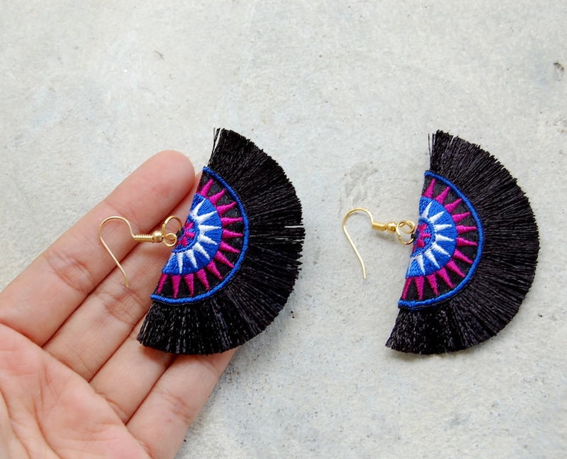 May include: A pair of black tassel earrings with a colorful geometric design. The earrings feature a blue, white, and pink mandala pattern.