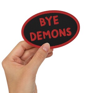 May include: An oval-shaped embroidered patch with a black center and a red border. The words "BYE DEMONS" are stitched in red across the center. The patch is held up by a hand.