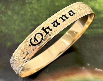 May include: Gold Hawaiian bangle bracelet with an engraved floral design and the word "Ohana" in black lettering.