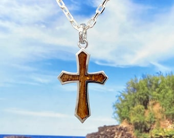 Op de afbeelding: Een zilveren ketting met een houten kruis hanger. Het kruis is gemaakt van koa-hout en heeft een zilveren rand. De ketting is gefotografeerd tegen een achtergrond van blauwe lucht en oceaan.