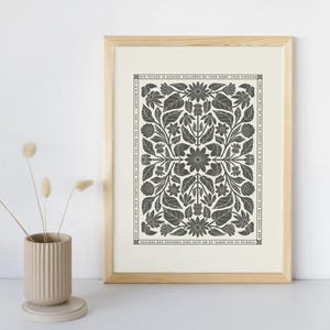 May include: A framed print featuring a symmetrical floral design in shades of gray and white. The artwork includes text from Matthew 6:9-13, framed within a light wooden frame. A beige ceramic vase with dried flowers is in the foreground.