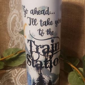 May include: White travel tumbler with a lid, featuring the text "Go ahead... I'll take you to the Train Station." The design includes a mountain scene with pine trees in shades of blue and black.