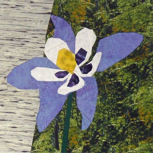 May include: A fabric art piece featuring a large, stylized blue and white columbine flower with a yellow center and dark purple accents. The flower is set against a backdrop of a forest scene and a birch tree.