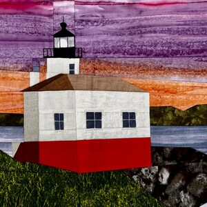 May include: A fabric art piece depicting a lighthouse on a rocky shore. The lighthouse has a red base, white walls, and a black top. The sky is a blend of purple, orange, and pink hues, suggesting a sunset or sunrise. The scene includes water and green grass.