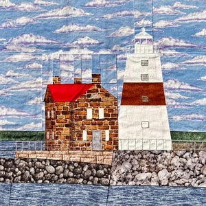 May include: A textile artwork showing a lighthouse and a building with a red roof. The lighthouse is white with a red band, and the building is made of brown bricks. The sky is blue with white clouds.