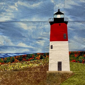 May include: A fabric quilt block depicting a lighthouse with a red top and white base, featuring a black lantern room. The scene includes a blue sky with white clouds, a dark blue horizon, and a foreground of brown, green, and autumn-colored foliage.