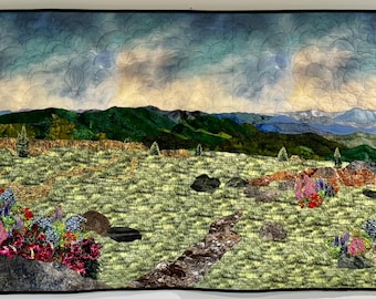 May include: A quilted wall hanging depicting a landscape scene with a mountain range in the distance, a field of wildflowers in the foreground, and a hot air balloon in the sky. The sky is a mix of blue and gray with clouds.