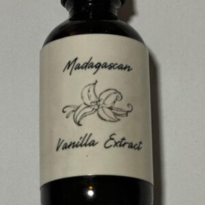 Könnte beinhalten: Eine dunkle Glasflasche mit Madagaskar-Vanilleextrakt. Das Etikett trägt die Aufschrift "Madagascan Vanilla Extract" mit einer Vanilleblüten-Illustration. Die Flasche hat einen schwarzen Deckel.