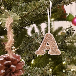 May include: A gold glitter letter A bauble hanging on a Christmas tree. The bauble is attached to a white ribbon.