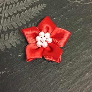 May include: A red satin flower with a white pearl center. The flower is on a dark gray background.