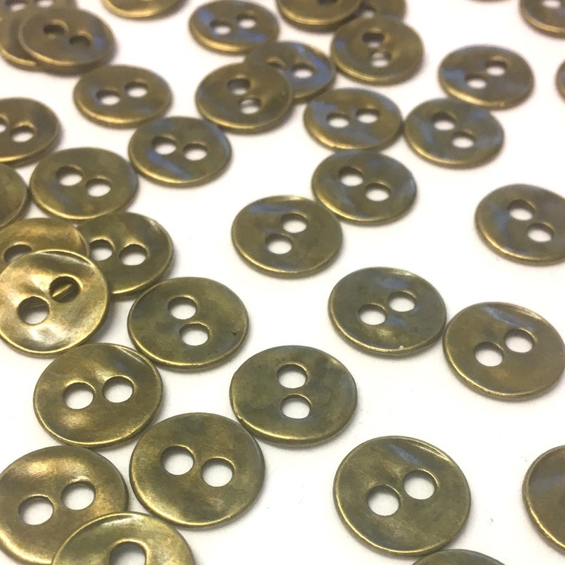 May include: A scattering of small, circular, brass-coloured buttons, each with two holes. The buttons are spread across a white surface, highlighting their metallic sheen and simple design. Suitable for sewing and craft projects.
