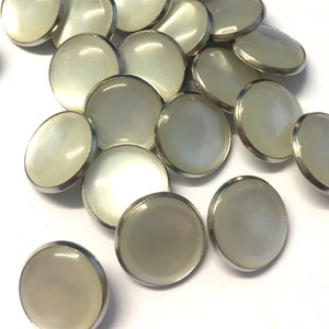 Könnte beinhalten: Eine Sammlung weißer runder Knöpfe mit silbernem Metallrand. Die Knöpfe sind auf einer weißen Oberfläche verstreut.