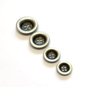 Pode incluir: Quatro botões de metal prateado com quatro furos cada. Os botões estão dispostos em uma fila do maior para o menor.