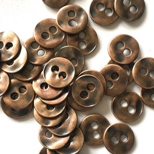 Könnte beinhalten: Eine Sammlung runder Knöpfe mit zwei Löchern und einem gealterten Kupferfinish. Die Knöpfe sind auf einer weißen Oberfläche verstreut und zeigen ihr gealtertes, metallisches Aussehen. Ideal für Näh- und Bastelprojekte.
