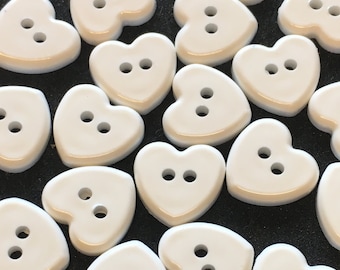 May include: A collection of white heart-shaped buttons with two holes each. The buttons are scattered on a black surface.