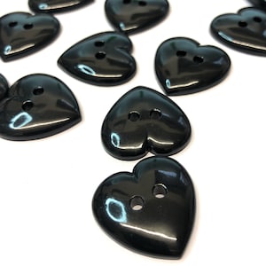 May include: A selection of glossy black heart-shaped buttons. Each button features two holes for sewing. The buttons are arranged on a white surface, with some overlapping. The buttons have a smooth, reflective finish.