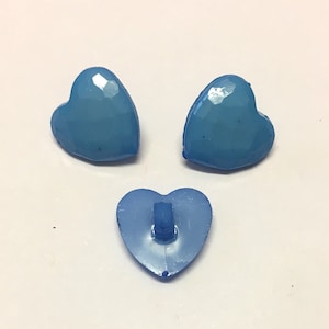 May include: Three light blue heart-shaped buttons. Two buttons have a faceted surface, while the third button shows the back with a loop for sewing. The buttons are a solid, opaque colour.