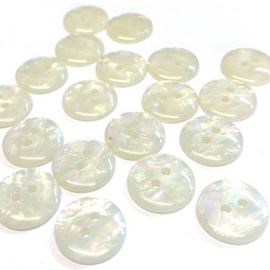 May include: A collection of iridescent, pearlescent buttons. Each button is round with two holes and a subtle, swirling pattern. The buttons have a creamy white colour and a glossy finish, suitable for sewing projects.