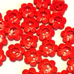 Könnte beinhalten: Eine Sammlung roter, plastikblumenförmiger Knöpfe mit jeweils zwei Löchern. Die Knöpfe sind in einem zufälligen Muster auf weißem Hintergrund angeordnet.