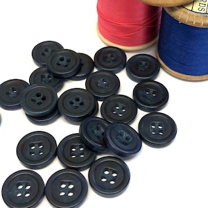 Könnte beinhalten: Eine Sammlung dunkelblauer Plastikknöpfe mit jeweils vier Löchern. Die Knöpfe sind auf einer weißen Oberfläche verstreut.