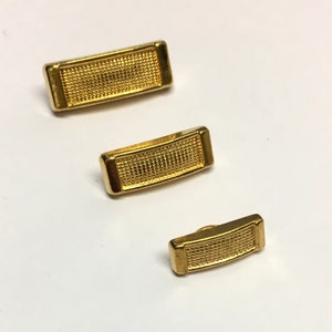May include: Three gold-coloured metal buttons with a textured surface. The buttons are rectangular and have rounded edges. The buttons are arranged in a row from largest to smallest.