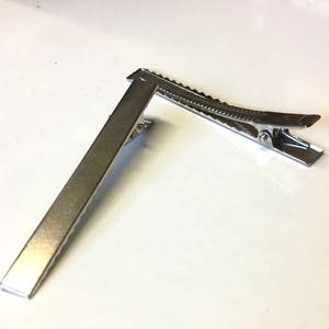Puede incluir: Una pinza para el cabello de metal plateado, con una base rectangular y una secci&oacute;n superior articulada y dentada. La pinza est&aacute; dise&ntilde;ada para sujetar el cabello. El metal tiene una superficie brillante y reflectante.