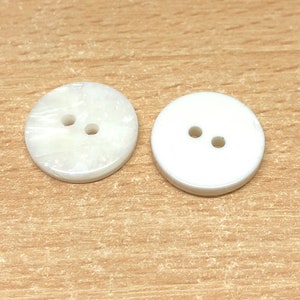 May include: Two white round buttons with two holes each. The button on the left has a textured surface, while the button on the right has a smooth surface.
