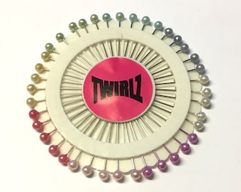 May include: A white plastic pincushion with a pink label that says "TWIRLZ". The pincushion has 48 colorful pins with iridescent glass heads.