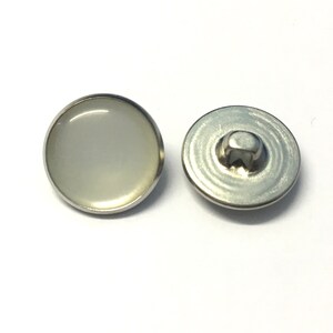 May include: A pair of white snap buttons with a silver metal rim. The button on the left has a white, opaque center. The button on the right has a silver metal back.