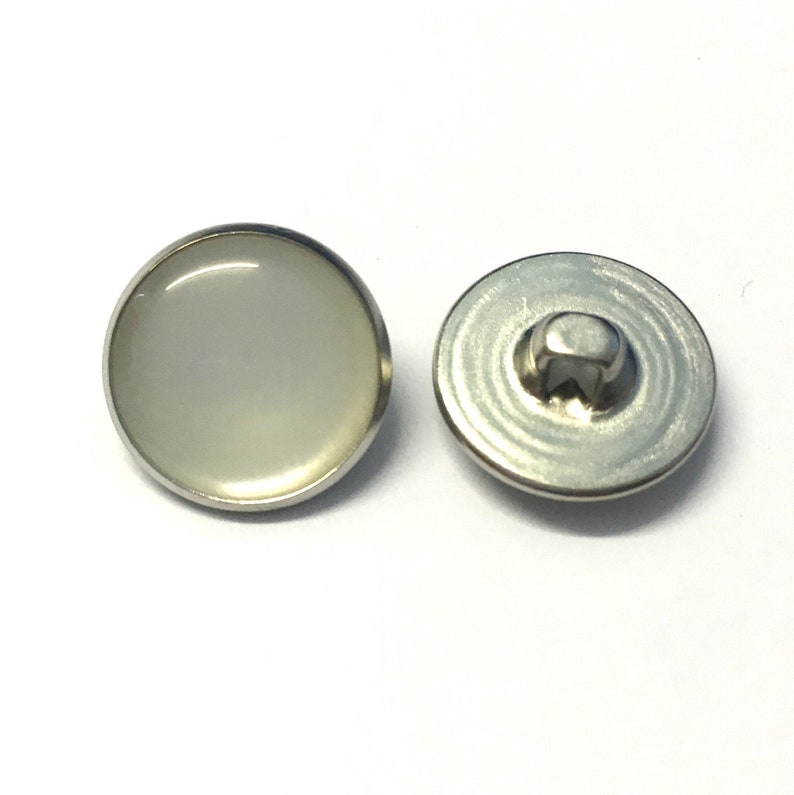 May include: A pair of white snap buttons with a silver metal rim. The button on the left has a white, opaque center. The button on the right has a silver metal back.