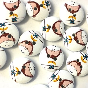 May include: A collection of 12 wooden buttons featuring a monkey design. The buttons are white with brown monkeys, yellow bananas, and blue flowers. Each button has two holes.