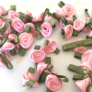 May include: A collection of small pink satin roses with green ribbon stems. The roses are arranged on a white surface.
