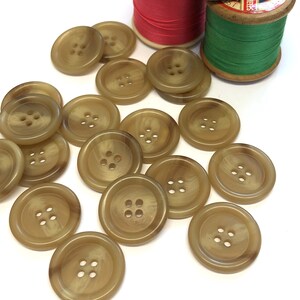 May include: A collection of large, round, tan buttons with four holes each, scattered on a white surface. Two spools of thread, one red and one green, are in the background. The buttons have a marbled appearance.