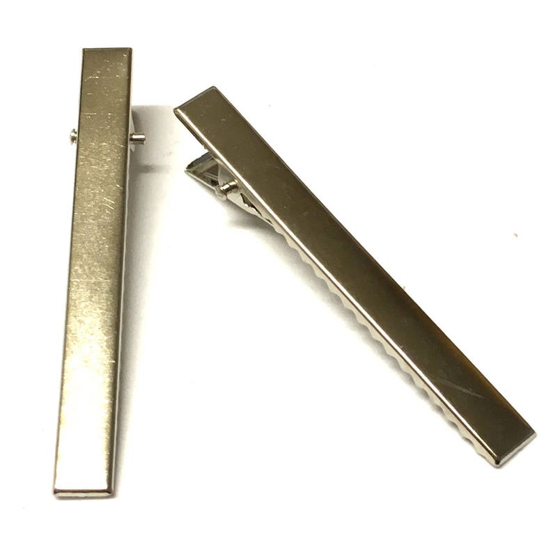 Puede incluir: Dos pinzas de pelo de metal plateado. Las pinzas son rectangulares con una parte superior plana y una parte inferior dentada con bisagras. Una pinza est&aacute; en posici&oacute;n vertical y la otra est&aacute; en &aacute;ngulo hacia la derecha, sobre un fondo blanco.