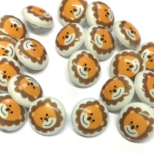 May include: A selection of round, white buttons featuring a cartoon lion face. The lion has an orange face, a brown mane, and a smiling expression. The buttons are made of a glossy material.