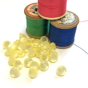 Może przedstawiać: Stosik jasnożółtych guzików z perłowym połyskiem. W tle znajdują się trzy szpulki nici, jedna czerwona, jedna zielona i jedna niebieska.