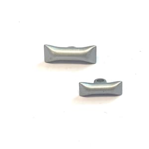 Puede incluir: Dos botones de metal plateado, uno con un bucle en la parte superior y el otro sin él. Ambos botones son rectangulares con bordes redondeados.