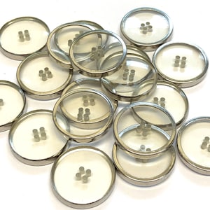 Puede incluir: Un grupo de 14 botones de plástico transparente con bordes de metal plateado. Cada botón tiene cuatro pequeños agujeros en el centro.