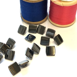 May include: A close-up of 15 black square buttons with a shiny finish. The buttons have a hole in the center for sewing.