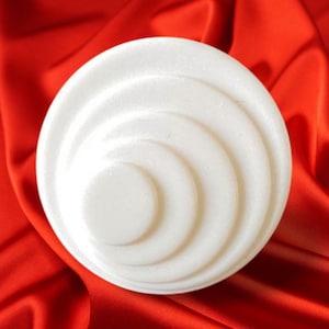 Pode incluir: Um objeto circular branco com um design concêntrico em camadas. O objeto está sobre um fundo vermelho vibrante e sedoso. O design apresenta uma série de anéis curvos e elevados, criando uma superfície texturizada. A estética geral é limpa e minimalista.