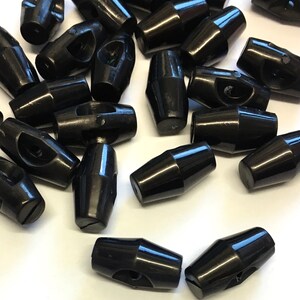 May include: A group of black plastic toggle buttons. The buttons have a hole in the centre and a small ridge on each side. They are commonly used for clothing and other crafts.